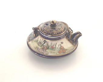 Może przedstawiać: Mały, ozdobny ceramiczny imbryk z niebiesko-złotym wzorem. Imbryk przedstawia malowaną scenę z postaciami w ogrodzie. Pokrywka ma złoty guziczek i pleciony uchwyt. Dzióbek i uchwyt są ciemnoniebieskie.