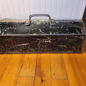 May include: A vintage metal toolbox with a black and green paint finish. The toolbox has a metal handle and is showing signs of wear and tear.