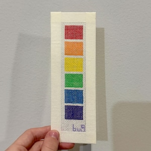 May include: A rectangular needlepoint bookmark featuring a rainbow color gradient of red, orange, yellow, green, blue, and indigo squares. The bottom of the bookmark has the initials "L.B.H." in blue. The bookmark is held against a neutral wall.