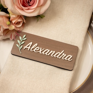 May include: A wooden place card with the name "Alexandra" in white script. A light green leaf design is on the left. The card rests on a beige napkin, with pink roses in the background.