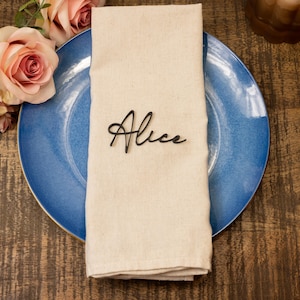 May include: A beige linen napkin with the name "Alice" in black script rests on a blue plate. Peach-coloured roses and greenery are arranged to the left. A candle holder is visible in the background. The scene suggests a table setting.