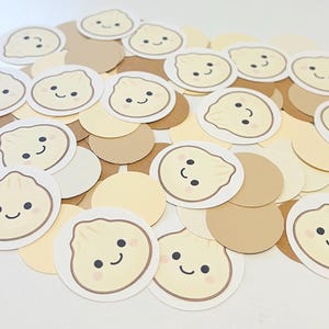 May include: A collection of circular paper confetti pieces in shades of cream, beige, and tan. Several pieces feature a printed illustration of a smiling dumpling with dark eyes, a small mouth, and pink cheeks, while others are solid coloured circles.