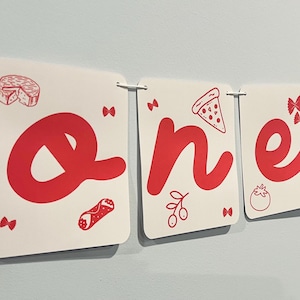 May include: A white banner consisting of three rectangular cards strung together on a white cord. Each card features a large red letter spelling out the word one, accompanied by red line-art illustrations of cheese, pizza, and pasta.