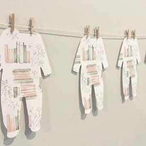May include: A decorative garland featuring multiple paper cutouts shaped like baby onesies, each suspended from a thin string by small wooden clothespins. The paper cutouts display a repeating pattern of stacked books and delicate floral sprigs.