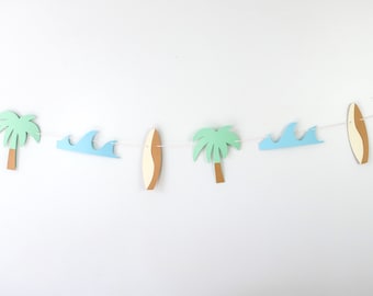 Puede incluir: Una guirnalda decorativa con formas recortadas de palmeras, tablas de surf y olas. Las palmeras tienen hojas de color verde claro y troncos marrones. Las tablas de surf son de color beige y blanco, y las olas son de color azul claro. La guirnalda está ensartada en una cuerda blanca.