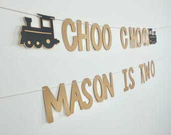 May include: Gold paper letters spelling "Choo Choo Mason is Two" with black paper train cutouts on a white background.