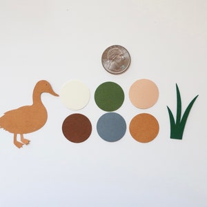 May include: A brown paper duck cutout with a white, brown, green, blue, and beige paper circles and a green paper grass cutout.