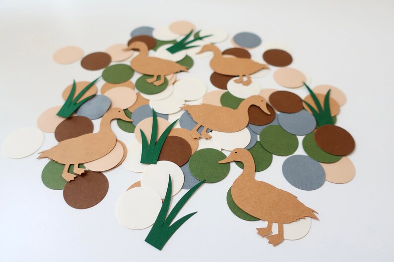 May include: Brown paper duck cutouts with green grass cutouts on a white background. There are also brown, beige, and gray paper circles.