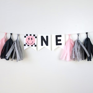 May include: Black and white banner with the word "ONE" in large black letters. The first letter has a pink smiley face with lightning bolts on a black and white checkered background. The banner is decorated with pink and black tissue paper tassels.