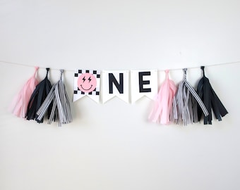 May include: Black and white banner with the word "ONE" in large black letters. The first letter has a pink smiley face with lightning bolts on a black and white checkered background. The banner is decorated with pink and black tissue paper tassels.