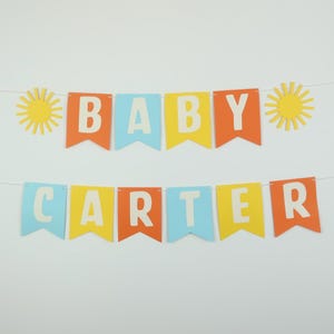 May include: A colorful banner with the words "BABY CARTER" in white letters on blue, yellow, and orange triangular flags. The banner is decorated with two yellow sun cutouts.