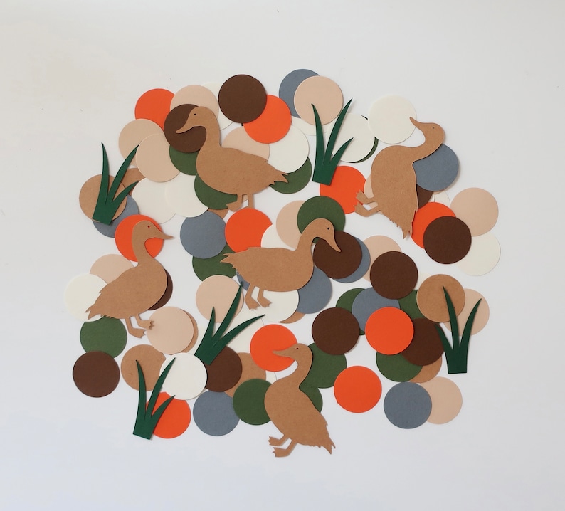 May include: A collection of brown paper ducks and green paper reeds on a white background surrounded by colorful paper circles in shades of brown, orange, white, and gray.