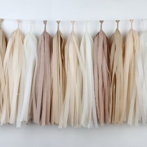 May include: A garland of paper tassels in various neutral colors, including beige, cream, and taupe, hangs against a white background. The tassels are made of thin, fringed paper strips, creating a textured, decorative element. Ideal for party decorations.