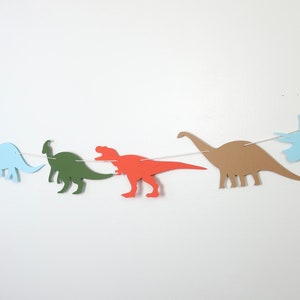 May include: A string of paper dinosaur cutouts in various colours, including blue, green, brown, and orange. The dinosaurs are strung together with white string and are hanging against a white wall.