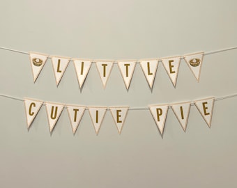 May include: A banner with the words "LITTLE CUTIE PIE" in gold letters. The pennants are white with gold trim and a pie graphic. The banner is strung on a thin white string, perfect for a baby shower or birthday party.