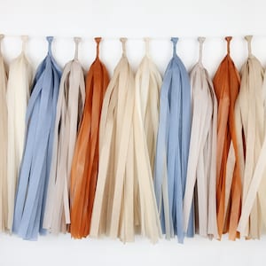 May include: A garland of tissue paper tassels in shades of beige, light blue, and orange. The tassels are strung together with white twine.