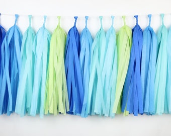 Puede incluir: Una guirnalda decorativa que presenta una serie de borlas de papel colgantes en tonos alternos de azul real, azul cielo, turquesa y verde lima. Cada borla individual está suspendida de un hilo blanco fino, creando una fila uniforme.