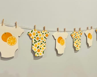 Könnte beinhalten: Eine Girlande aus Baby-Bodys, die mit Holzklammern an einer Schnur befestigt sind. Die Bodys sind mit Orangen- und Zitronen-Illustrationen verziert. Der Hintergrund ist eine schlichte, hellgraue Wand.