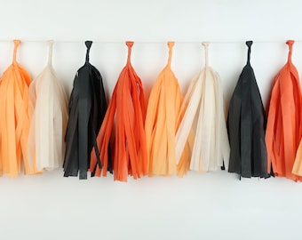 May include: A string of tissue paper tassels in orange, black and cream colours. The tassels are hanging from a white string.