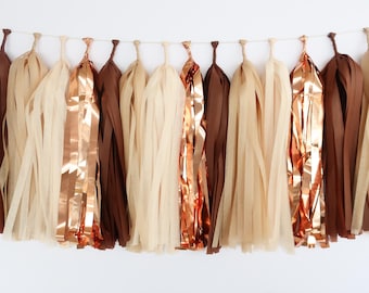May include: A decorative garland of paper tassels in shades of brown, beige, and copper. The tassels are suspended from a thin string, creating a festive and elegant look. Ideal for party decorations or home decor.