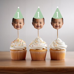 Peut inclure: Trois cupcakes avec un gla&ccedil;age blanc et des p&eacute;pites color&eacute;es surmont&eacute;s d'un chapeau de f&ecirc;te vert et blanc avec le chiffre "1" et une photo d'un b&eacute;b&eacute; portant le m&ecirc;me chapeau.
