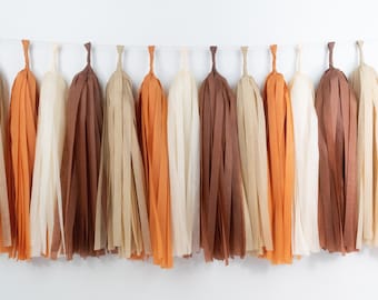 May include: A decorative garland of paper tassels in shades of beige, orange, and brown. The tassels are suspended from a thin white string, creating a festive and colorful display. The tassels are made of thin strips of paper.