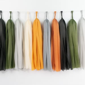 May include: A colorful garland of paper tassels in shades of gray, orange, black, green, and cream. The tassels are suspended from a white string, creating a festive and decorative display. Perfect for parties and celebrations.