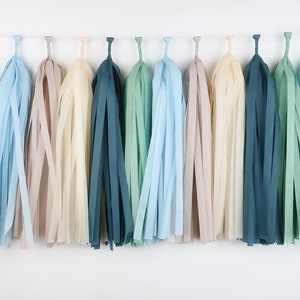May include: A garland of tissue paper tassels in shades of blue, green, and beige. The tassels are strung together on a white string and are hanging from a white background.