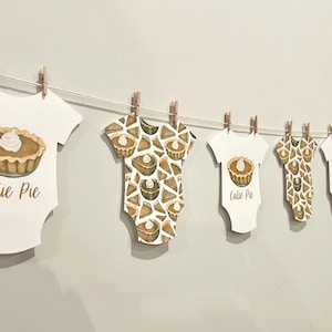 May include: A string of baby onesies, each clipped to a clothesline with wooden clothespins. The onesies are white with a pie graphic and the words "Cutie Pie". Some have a pie pattern.