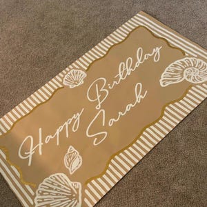 May include: A rectangular sign with a tan background and white text that reads "Happy Birthday Sarah." The sign is decorated with white seashell illustrations and a gold border with white stripes.