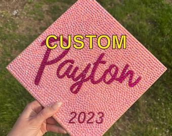 May include: A pink graduation cap adorned with tiny, shimmering rhinestones. The cap displays the word "CUSTOM" in yellow block letters, the name "Payton" in a cursive, magenta font, and the year "2023" in a similar magenta font.