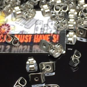 May include: A close-up of a pile of silver earring backs. The backs are made of metal and have a small hole in the center. They are designed to be used with post earrings.