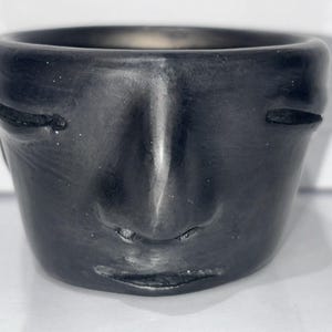 May include: A black ceramic face planter with a detailed facial design. The planter has a rounded shape with a wide opening and a sculpted face on the front. The face features closed eyes, a nose, and a smiling mouth.