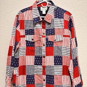 May include: A patchwork jacket in red, white, and blue, featuring squares of stripes, stars, and gingham patterns. The jacket has a collared neckline, button-down front, and two chest pockets. It is hanging on a white hanger.