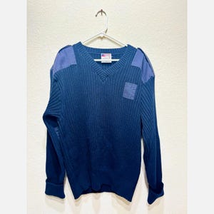 May include: Navy blue V-neck sweater with ribbed knit texture. Features shoulder patches and a small square patch on the chest. The sweater is hanging on a white hanger against a white background. The label is visible.