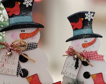 May include: Two white wooden snowman decorations featuring black top hats with red cardinal birds and white snowflakes. Each snowman wears a checkered scarf, has a carrot nose, and holds a tag with the text Let it Snow and a small bell.