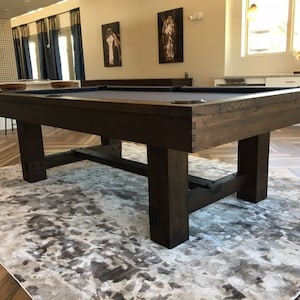 May include: A dark brown wooden pool table with a dark green felt top. The table has a rustic, reclaimed wood look and is set on a light gray rug.