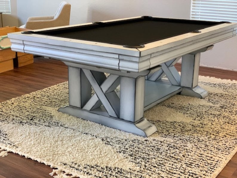 May include: A white distressed pool table with a black felt top and a wooden X-shaped base. The table is on a white rug.