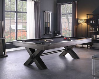 May include: A black pool table with a dark wood finish and a black felt top. The table has an X-shaped base with a metal bar connecting the two sides. The table is set up in a game room with a window in the background.