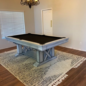 May include: A white pool table with a black felt top and a distressed wood finish. The table is set up in a room with wood floors and a white rug.