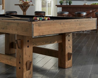 May include: A rustic wooden pool table with a black felt playing surface and a set of billiard balls. The table has a simple, sturdy design with thick wooden legs and a wide, flat top.