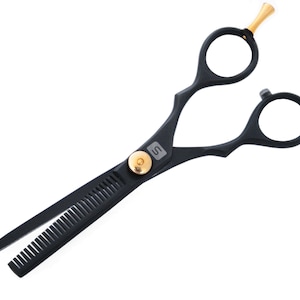Puede incluir: Tijeras de peluquería negras con detalles dorados y un mango negro texturizado. Las tijeras tienen una cuchilla dentada en un lado.