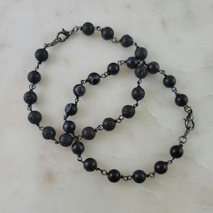 May include: Two black beaded bracelets with a dark metal clasp. The bracelets feature a mix of matte black beads, textured lava rock beads, and patterned agate beads, strung on a thin, dark wire.