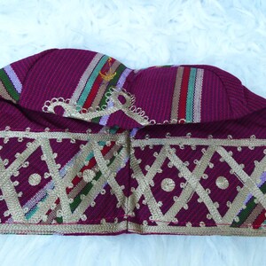 May include: A burgundy fabric hat with gold geometric embroidery. The hat features vertical stripes in red, green, and white. The hat is likely a cultural or ceremonial head covering.