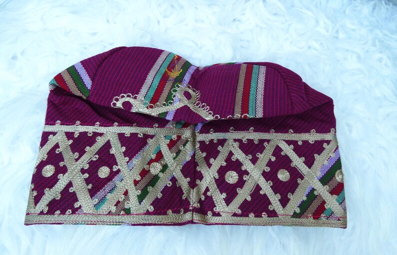 May include: A burgundy fabric hat with gold geometric embroidery. The hat features vertical stripes in red, green, and white. The hat is likely a cultural or ceremonial head covering.