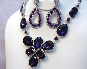 May include: A silver-toned jewelry set featuring a necklace and matching earrings. The necklace and earrings are adorned with large, dark purple teardrop-shaped stones and smaller purple accents. The earrings are teardrop-shaped with a halo of smaller stones.