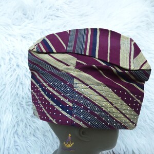 May include: A patterned cap in shades of burgundy, gold, and navy blue. The cap features stripes, diagonal lines, and dotted patterns. The fabric appears to be woven, with a slightly textured appearance. The cap is designed to fit the head.