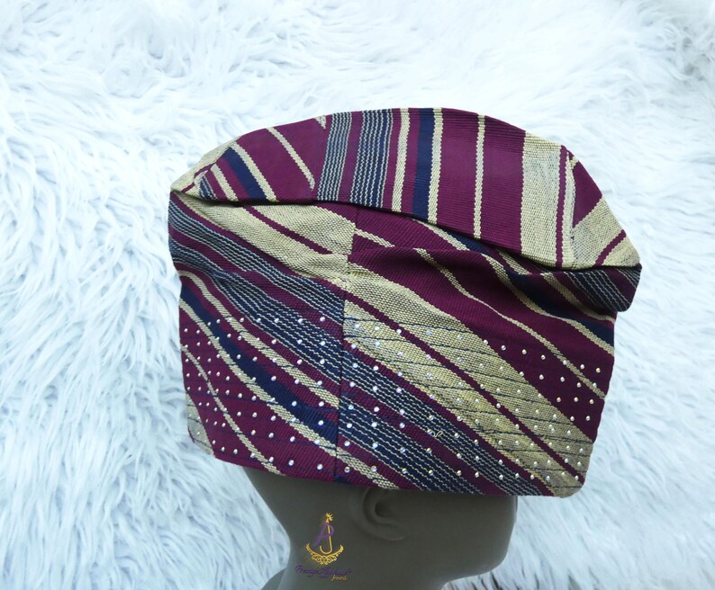 May include: A patterned cap in shades of burgundy, gold, and navy blue. The cap features stripes, diagonal lines, and dotted patterns. The fabric appears to be woven, with a slightly textured appearance. The cap is designed to fit the head.