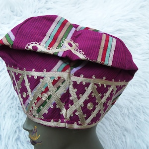 May include: A magenta-colored hat with a patterned design. The hat features gold embroidery and colorful stripes. The hat is likely a cultural or ceremonial head covering.