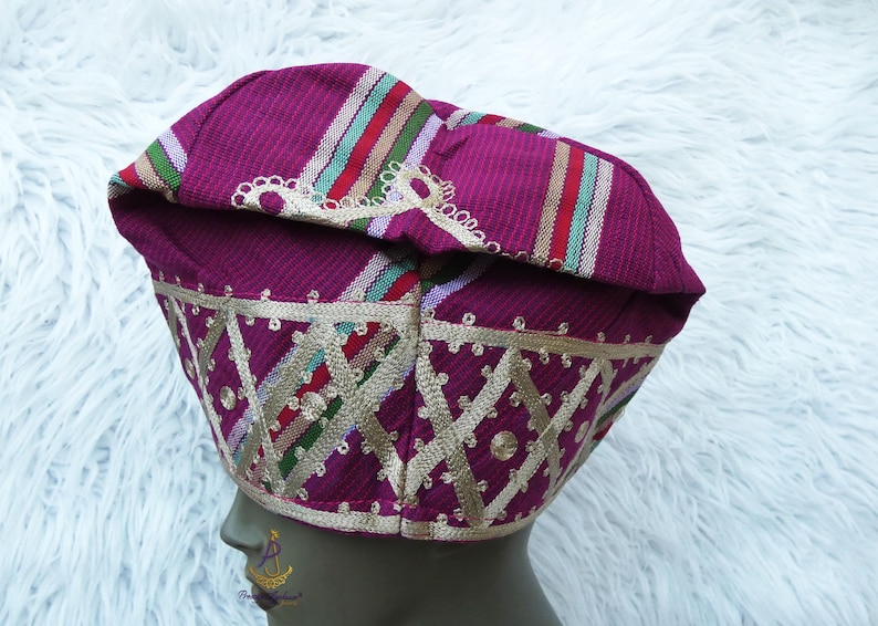 May include: A magenta-colored hat with a patterned design. The hat features gold embroidery and colorful stripes. The hat is likely a cultural or ceremonial head covering.